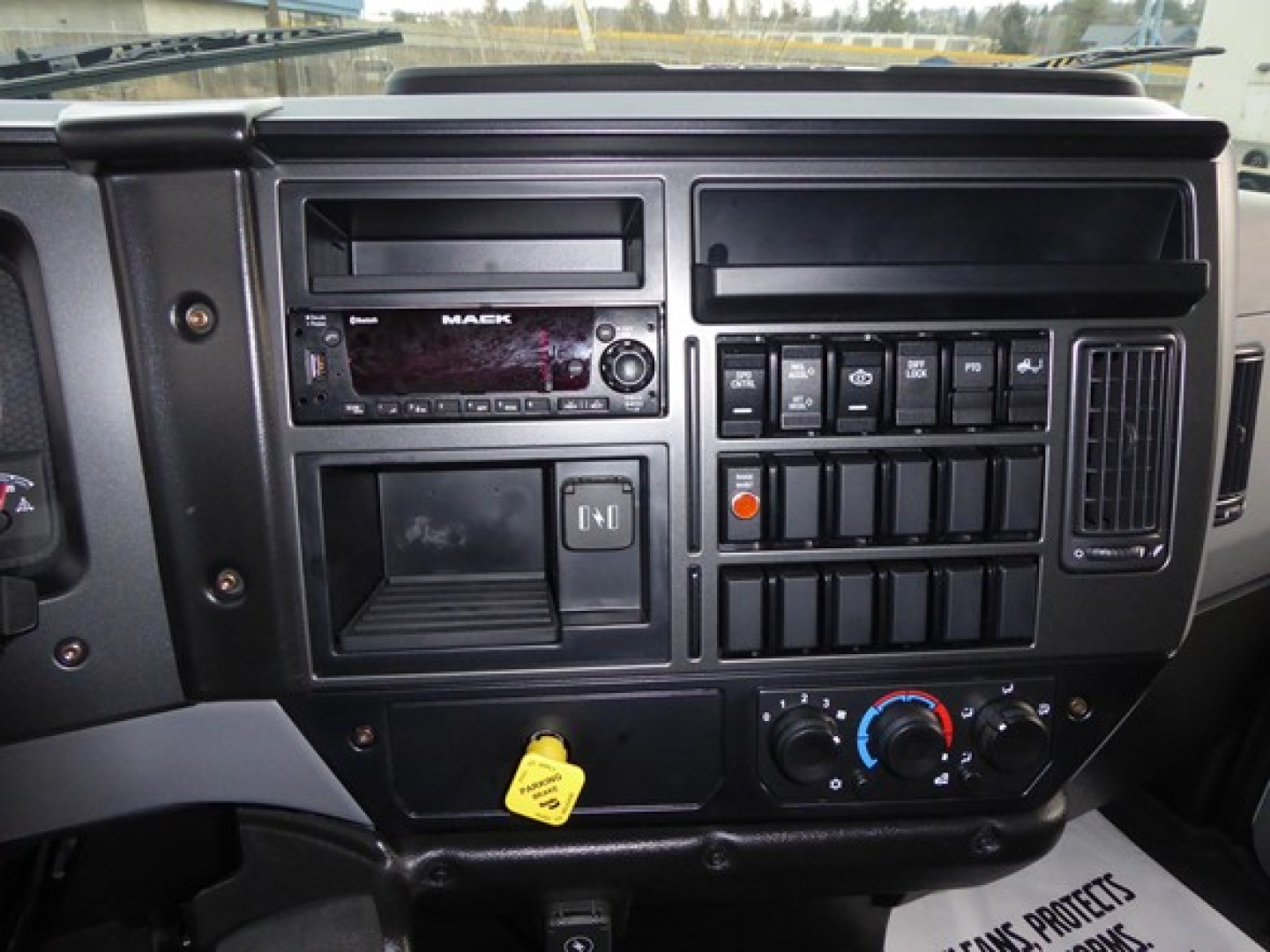 Truck dashboard showing Mack stereo, control switches, climate knobs, ignition yellow key tag in lower left.