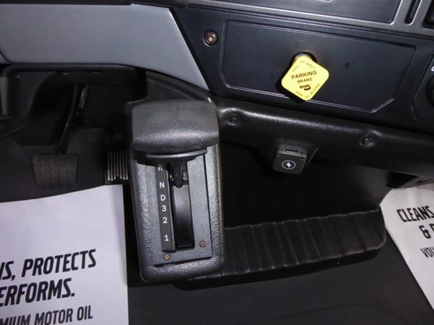 Gear selector with PRND32 indicator in Mack MD6 cab showing gear positions. Nearby dashboard switch and a yellow parking brake warning label. Floor mat visible.