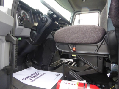 Interior cab view of a 2025 Mack MD6 truck showing driver’s seat, steering wheel, and dashboard with pedals; floor mat with text; part of passenger area.