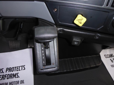Gear selector with PRND32 indicator in Mack MD6 cab showing gear positions. Nearby dashboard switch and a yellow parking brake warning label. Floor mat visible.