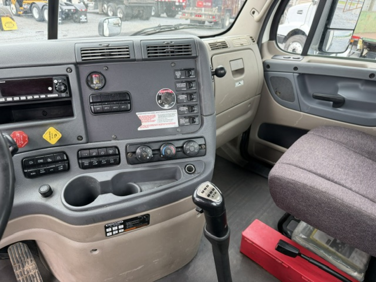 Dashboard and driver-side cab interior of a Freightliner Cascadia showing control panel, gear shifter, cup holders, and window controls.