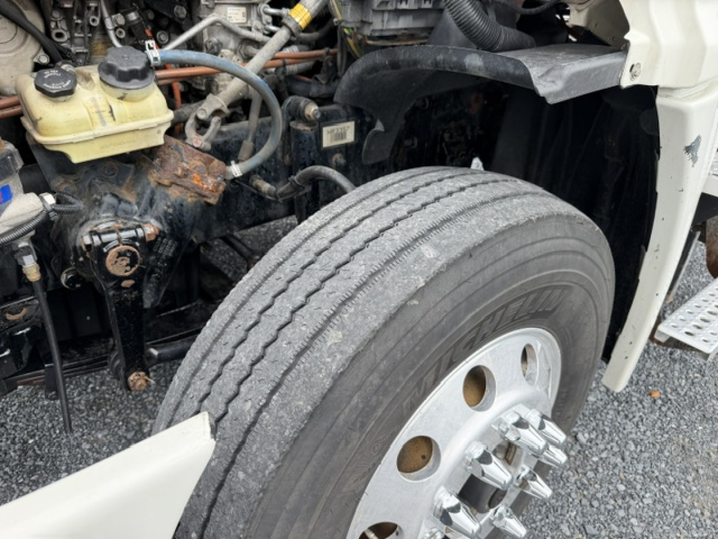 Truck engine bay with front left wheel visible; various hoses, a brake fluid reservoir, and engine components. No legible model text observed.