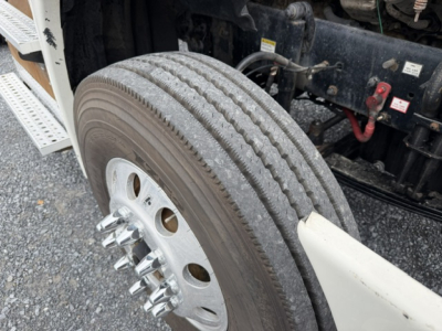 Truck front wheel and chassis of a Freightliner Cascadia daycab with tire tread and wheel nuts visible; partial undercarriage components and a red connector visible.