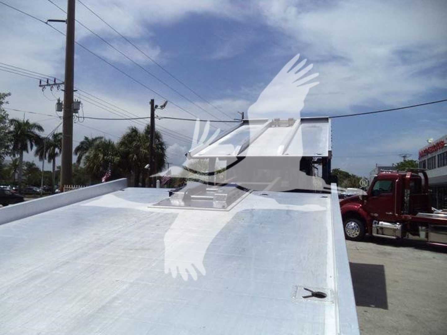 Flatbed tow truck bed with chrome metal surface and a white hutch cab in the background; windshield and front of another truck visible.