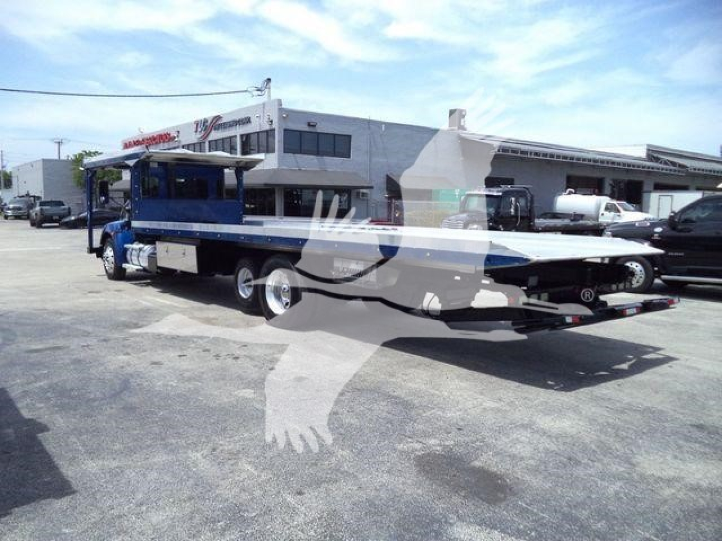 Kenworth T370 rollback truck with flatbed and blue cabin, side view showing the trailer and hydraulic system under a clear sky.