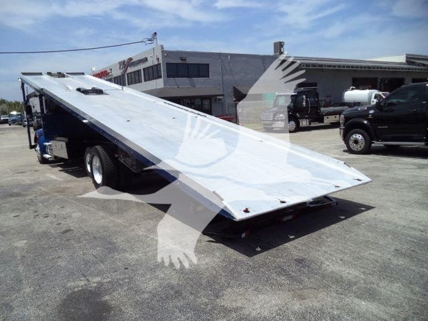 Kenworth T370 rollback truck with raised flatbed in a dealership lot, multiple vehicles in background.