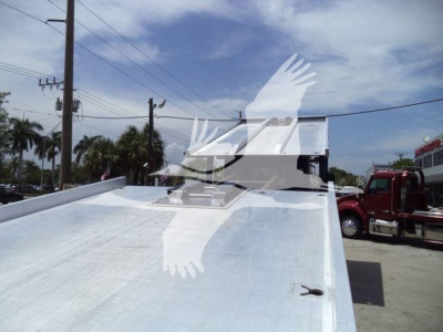 Flatbed tow truck bed with chrome metal surface and a white hutch cab in the background; windshield and front of another truck visible.