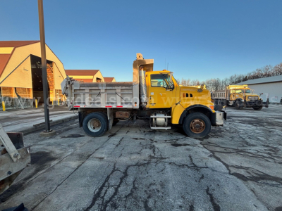 Dump truck side view showing yellow Sterling L8500 chassis with dumping bed, front cab, black tires, and blue sky.