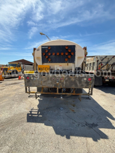 Rear view of a tanker truck trailer with multiple orange hoses and a 'WATER' placard, showing a circular tank and metal bumper.
