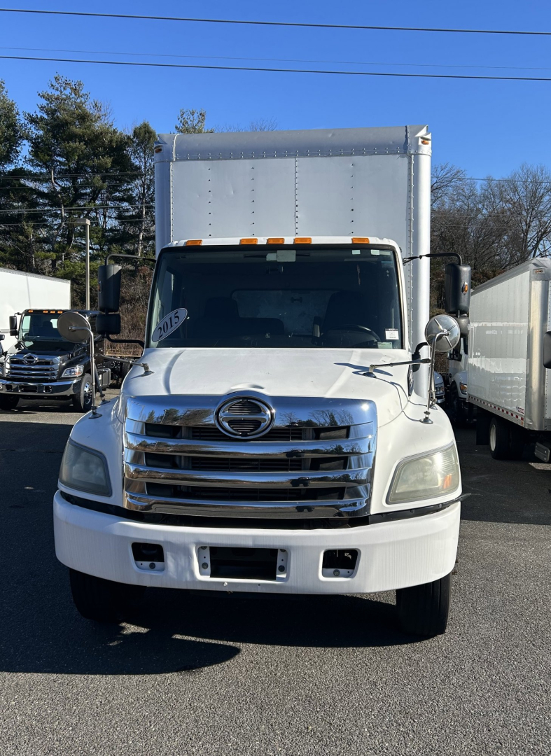 Front view of a white 2015 Hino 338 box truck with large grille, Hino emblem, horizontal chrome bars, rectangular headlights, and a box cargo area behind the cab.