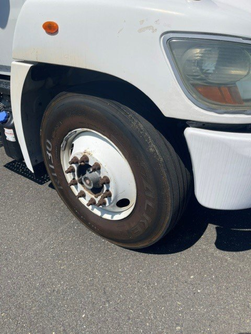 Front wheel and fender of a white 2015 Hino 338 box truck showing wheel, tire, and headlight area with partial fender flare.