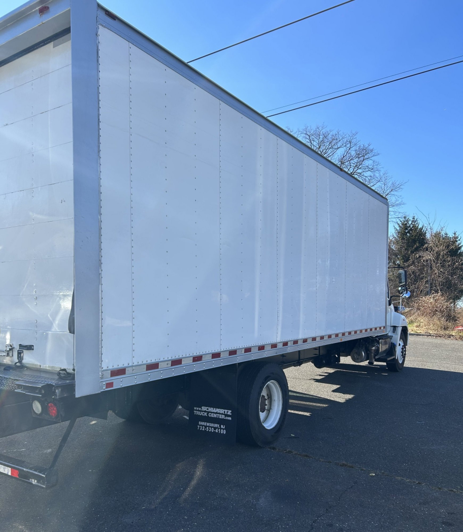 Box van truck in profile view showing large rectangular cargo box on chassis with white exterior, rear and side rails, single rear wheel, and a cab in the background.