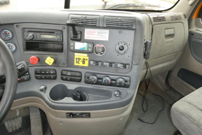 Truck cab interior showing dashboard gauges, controls, and center console of a Freightliner Cascadia Daycab; visible labels include a yellow '12-6' tag and various switches.
