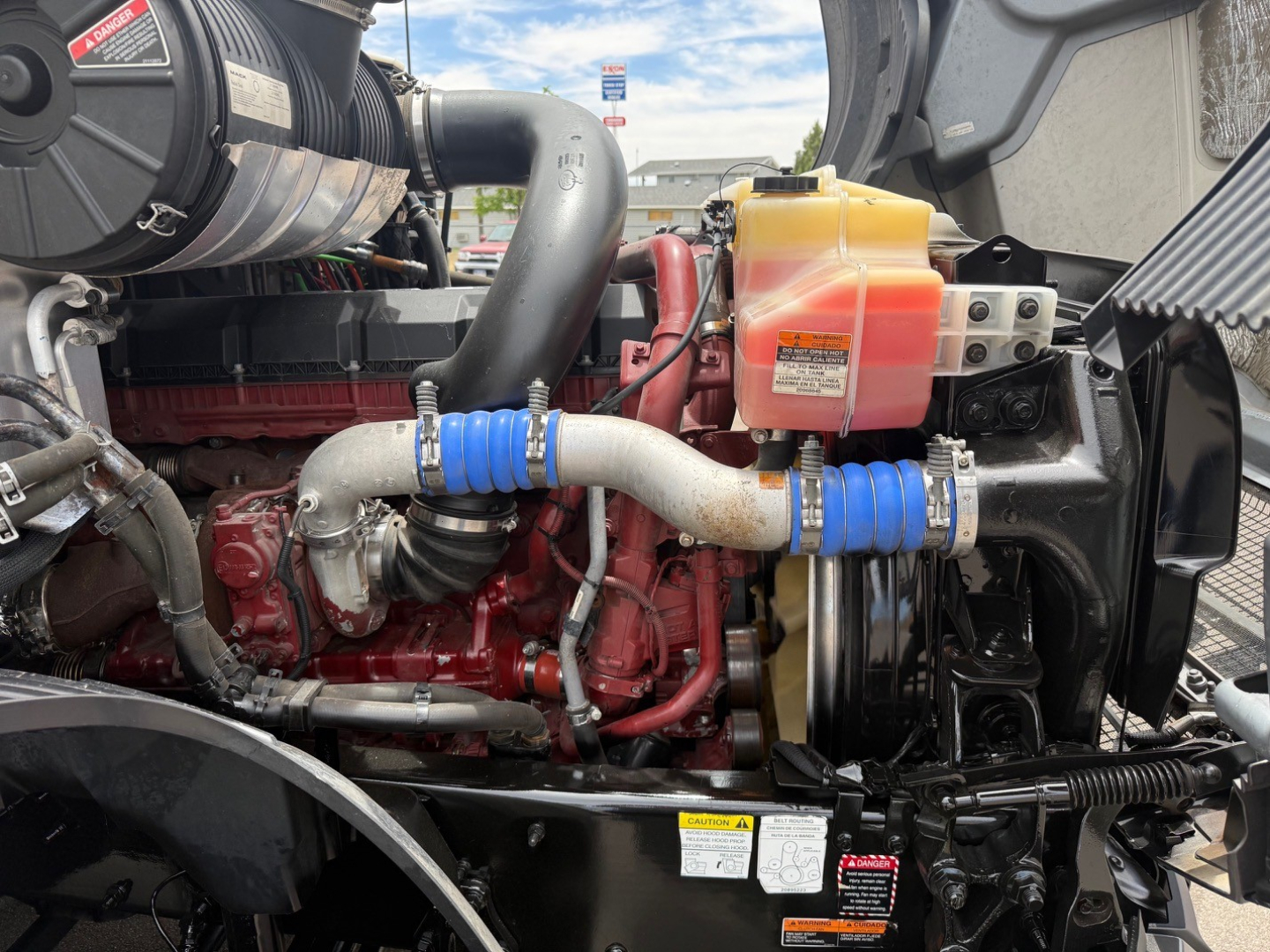 Truck engine bay showing coolant reservoir with pink/orange coolant, blue hose clamps, intake piping, and red engine block visible.
