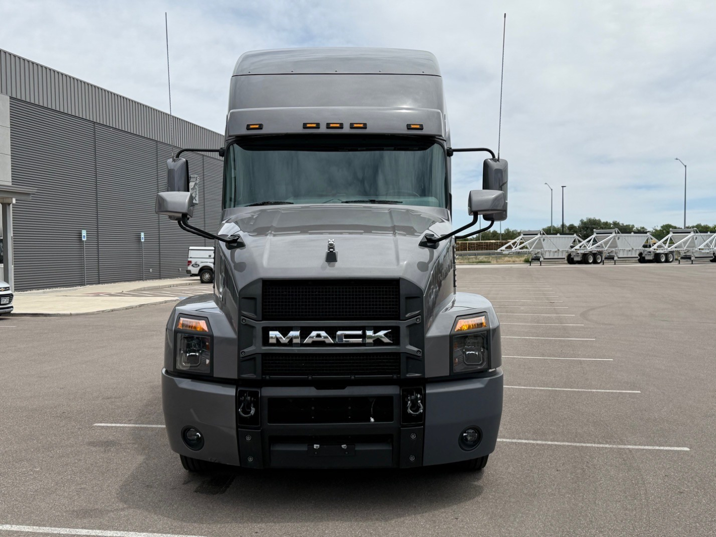 Front view of a Mack truck with a large black grille and 'MACK' logo, headlights, and grey cab in a parking lot.