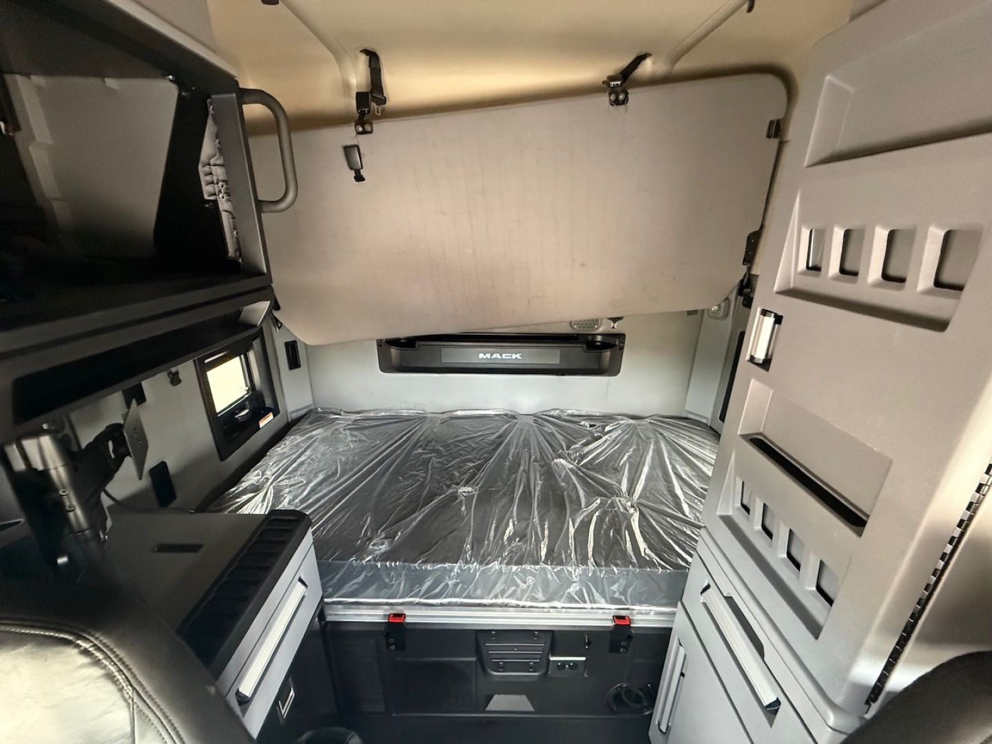 Truck cab sleeper interior with a platform bed in the rear wall, side storage, and control panels. Visible text includes MAHEN or MA KOM? on the bed shield and a small label on the dashboard area.