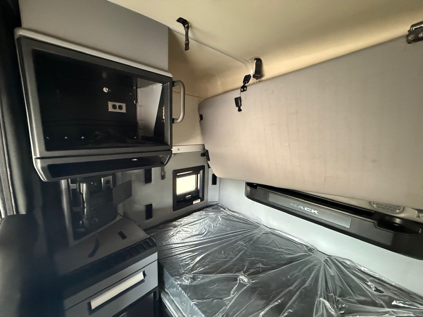 Truck cab interior showing overhead storage and sleeper area with plastic-covered mattress, light grey panels, and control compartments.