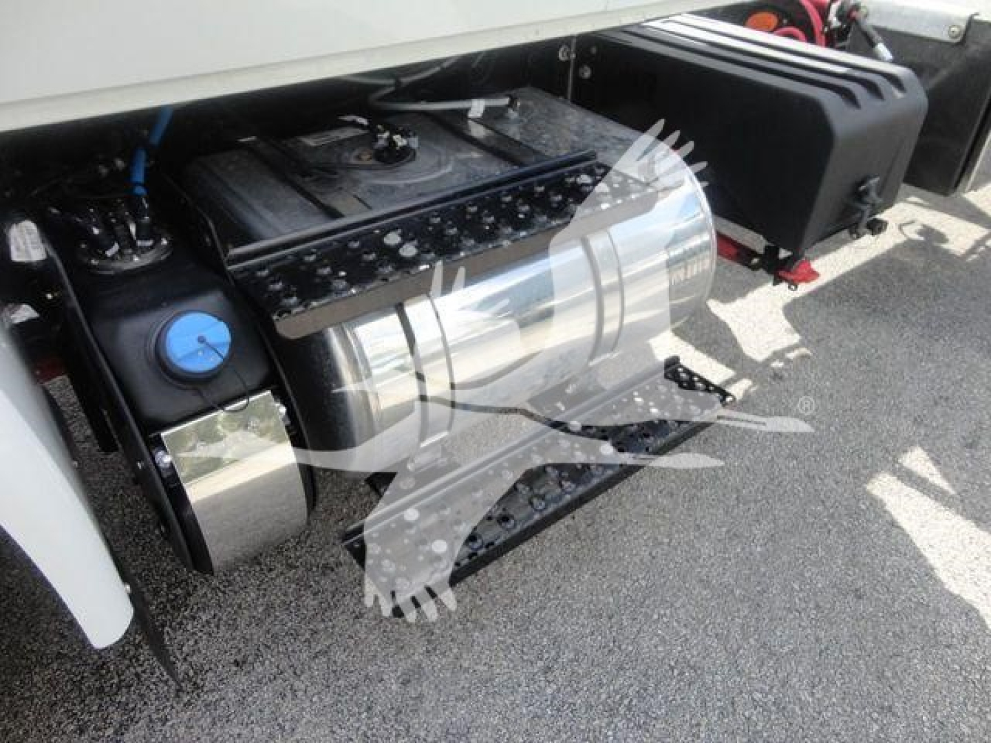 Truck fuel tank and undercarriage view showing cylindrical stainless fuel tank, blue cap, and nearby black equipment; part of engine bay/drive train area visible.