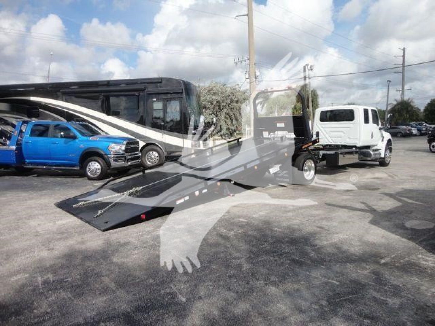 International MV tow truck with a ramp-loading bed and a white truck in the pickup area; multiple vehicles in background.