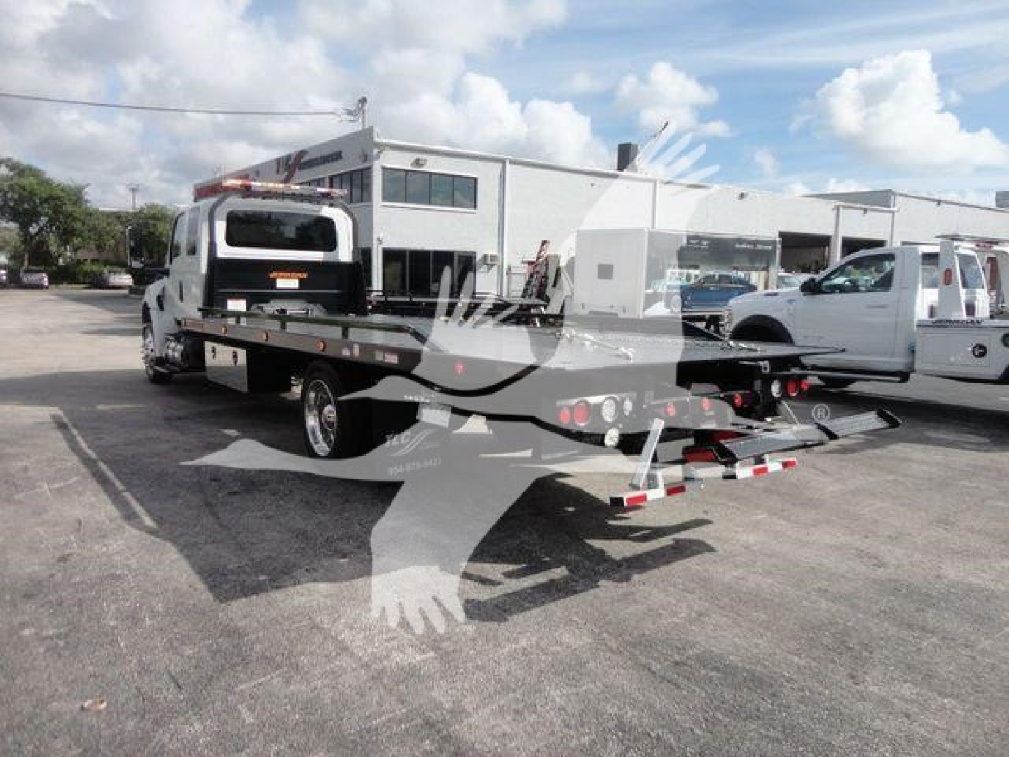 Tow truck with rollback bed attached, International MV model, white cab, in a dealership lot.