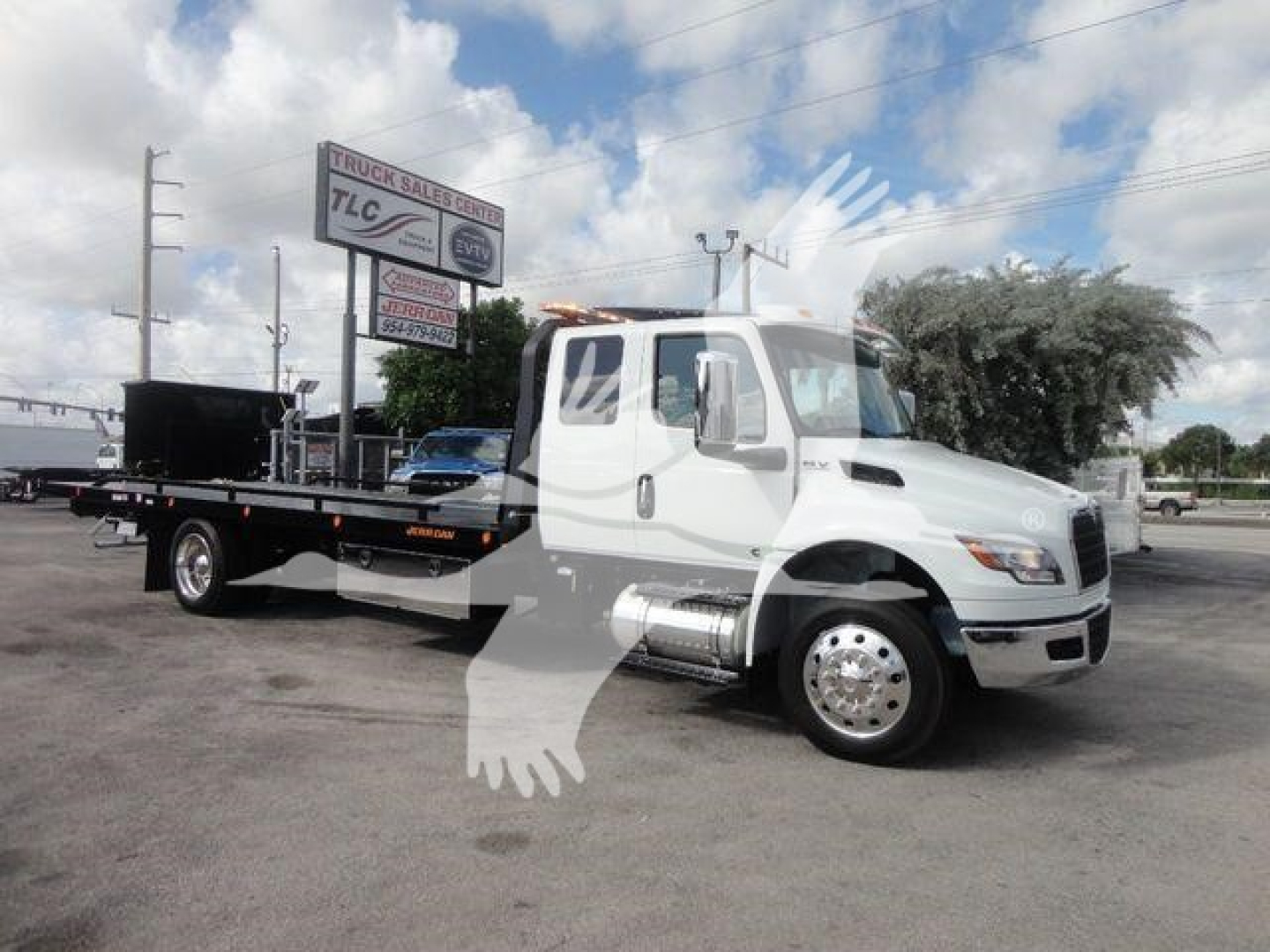 Truck cab and flatbed rollback tow truck showing white International MV with extended cab and long flatbed, stock number MV607EXCAB22XLP, dealership in background.