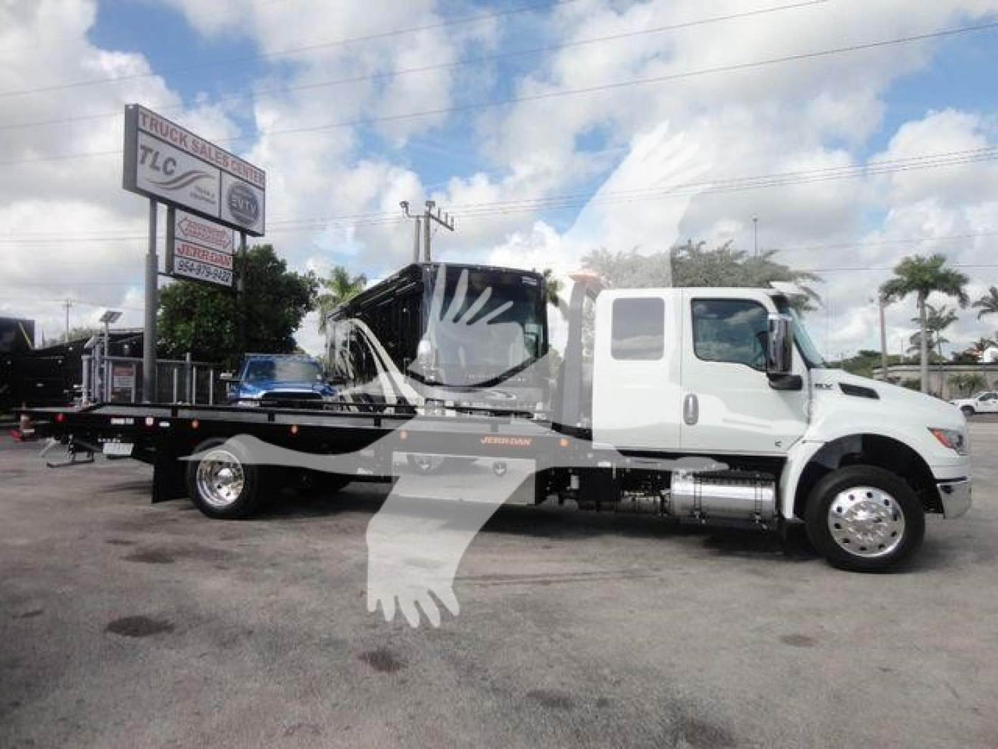 Tractor-trailer truck with a white cab and a flatbed rollback bed carrying a damaged vehicle. Misc signage in background.