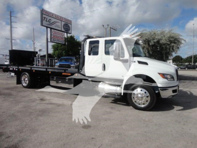 International MV flatbed tow truck in white cab with rollback bed, stock image showing side profile and dealership sign in background.
