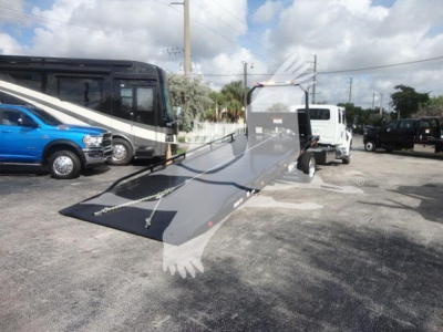 Truck with a ramp in a parking lot showing a white tow truck and a black and white bus in the background; MV607EXCAB22XLP text not visible.