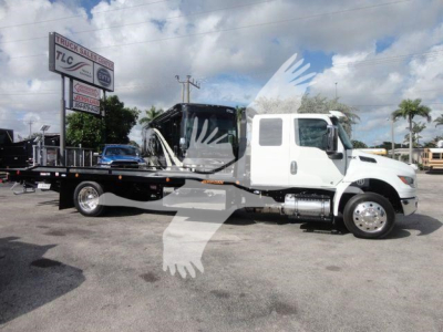 Truck showing a white International MV rollback wrecker body with a long flatbed and a tow boom. Visible text on signs and dealership in background.