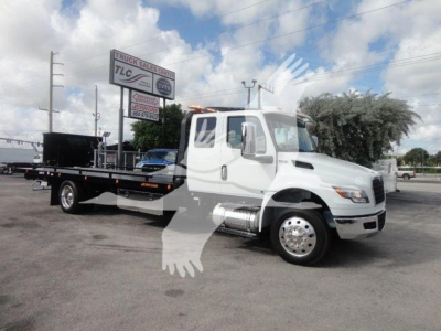Truck cab and flatbed rollback tow truck showing white International MV with extended cab and long flatbed, stock number MV607EXCAB22XLP, dealership in background.