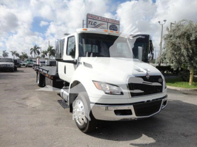 Truck bay showing a white 2027 International MV rollback truck with a flatbed. Visible text on sign reads TRUCK SALES CENTER and TLC.