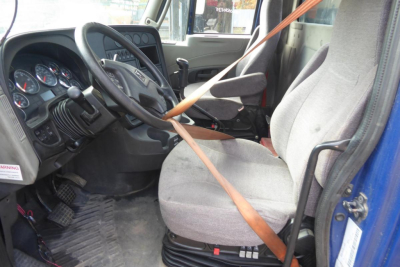 Int'l Prostar cab interior showing steering wheel, dashboard gauges, and driver seat with seat belt across. Visible pedals and entry area.