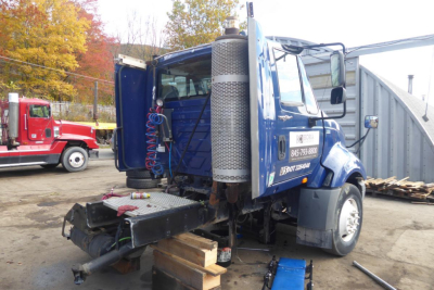 Semi truck cab exterior showing blue International ProStar+ Daycab with open rear doors, vertical exhaust stack, and tools around the chassis.