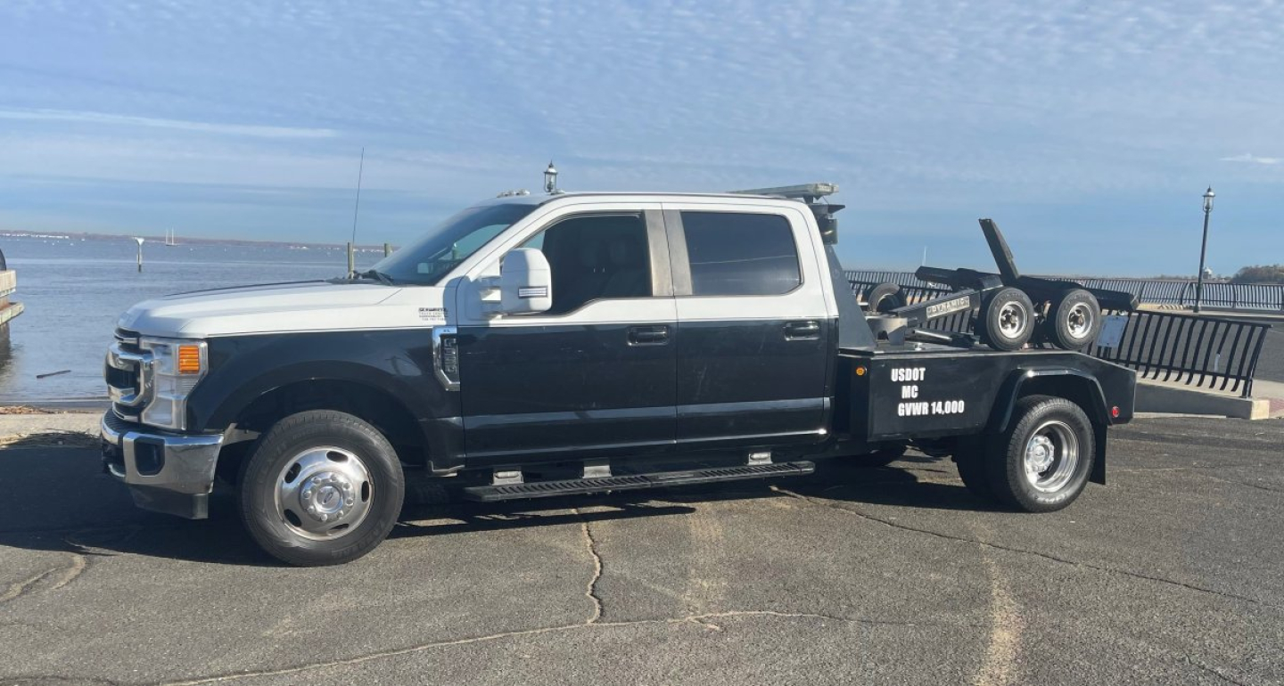 Truck towing a flatbed tow truck with dual rear wheels and equipment on bed, white cab and black flatbed, Ford emblem visible, harbor background.