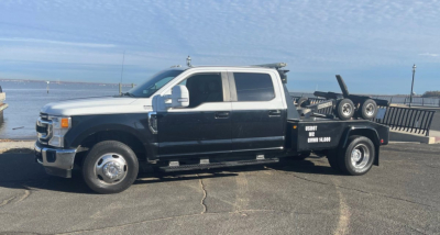 Truck towing a flatbed tow truck with dual rear wheels and equipment on bed, white cab and black flatbed, Ford emblem visible, harbor background.