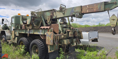 AM General M816 wrecker truck with boom crane and weathered military camo, side view showing crane arm elevated with hook and stabilizers.