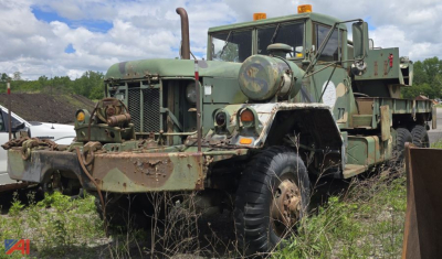 AM General M816 wrecked wrecker truck in-service showing front bumper winch and boom lift attachment, green military camouflage with dirt and overgrown grass.