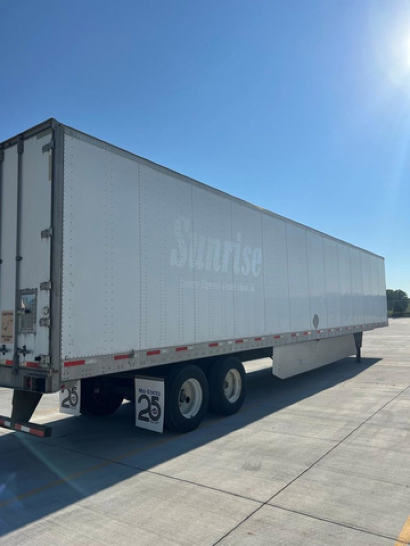 Semi-trailer/box trailer showing 'Sunrise' branding along the side, tandem rear wheels, and a 25 mph sign mounted under the trailer near the back. Clear blue sky.