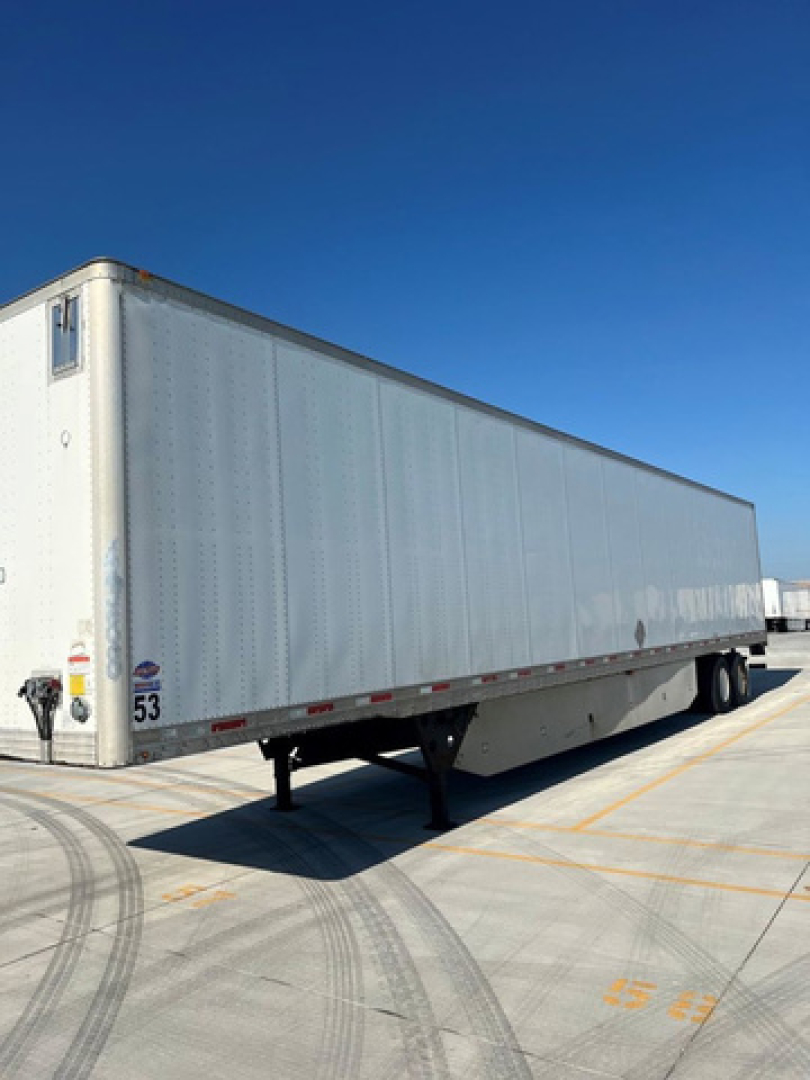 Trailer van side view showing aluminum box trailer with landing gear and wheel axles, numbered 53 near rear, under clear blue sky.