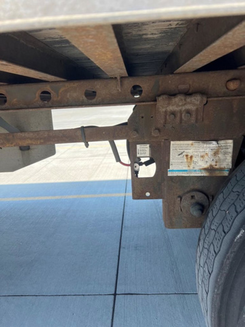 Trailer undercarriage showing rusted frame rails with a wheel and a small label, utility van trailer type, visible riveted metal and mounting bracket.