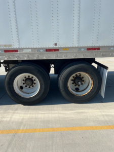 Dual trailer wheels visible under a silver cargo trailer with riveted side panels and reflection on the concrete.