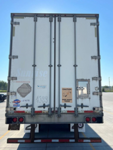 Rear view of a white utility van trailer with double doors, metal latches, and warning labels; VIN and stock stickers faintly visible on doors.