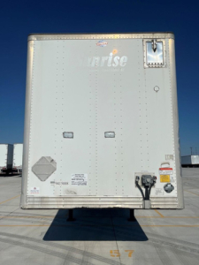 Rear view of a light-colored utility trailer with 'unrise' branding and rear door, showing safety decals and hitch components.