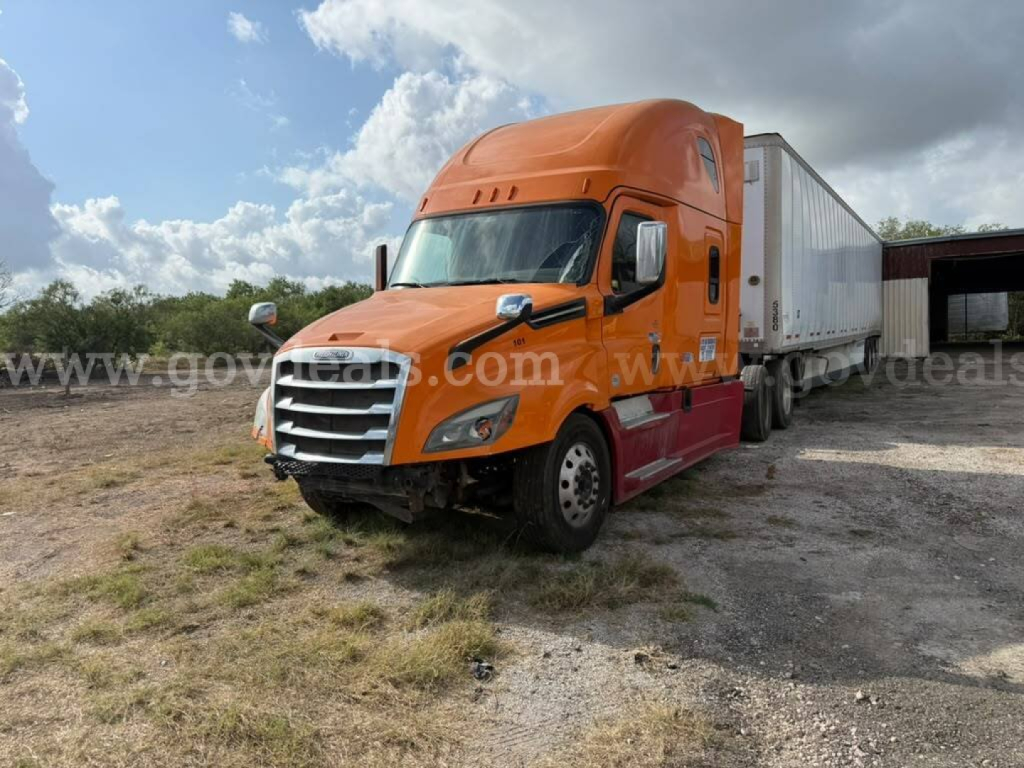 Freightliner Cascadia truck in orange color with a long trailer attached, parked on a dirt lot.