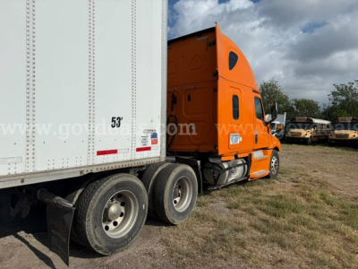 Truck cab and trailer from a 2020 Freightliner Cascadia with orange cab connected to a white trailer, dual rear axles visible, yard with other trucks in background.