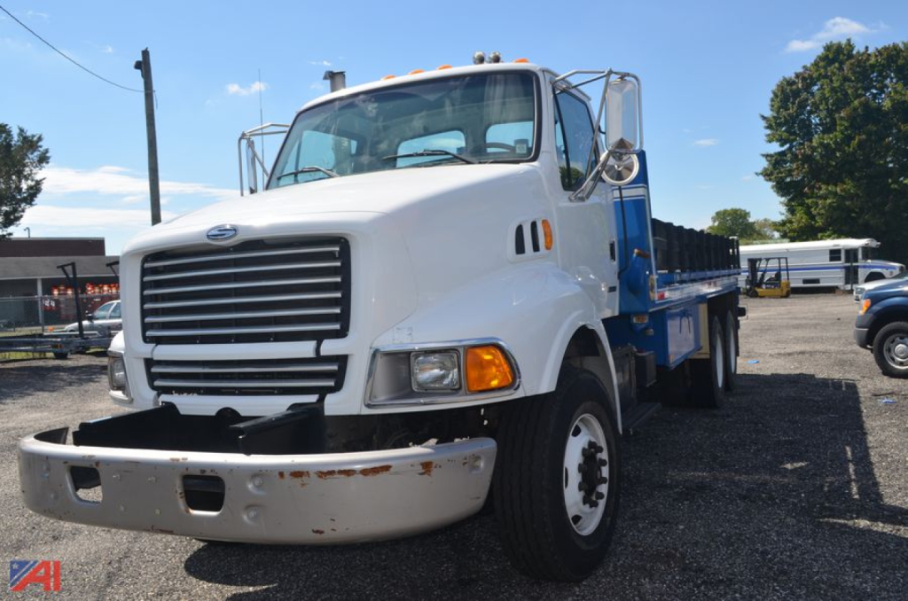 Flatbed truck front-left view showing white cab with large grille and bumper, Sterling L8500 series badge not visible, blue flatbed and side rails, daylight outdoor lot
