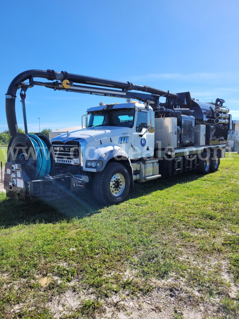 Vacuum truck with long hose reel and crane arm, white cab, multiple tanks rear, equipment on bed, clear blue sky.