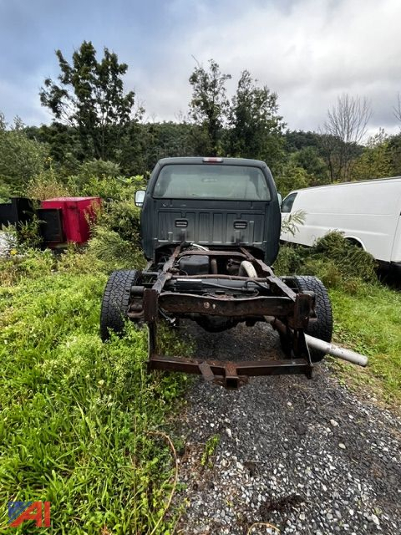 Rear view of a Ford F-350 chassis on a gravel lot with exposed frame and axles, green background, white van to the right, red container to left.