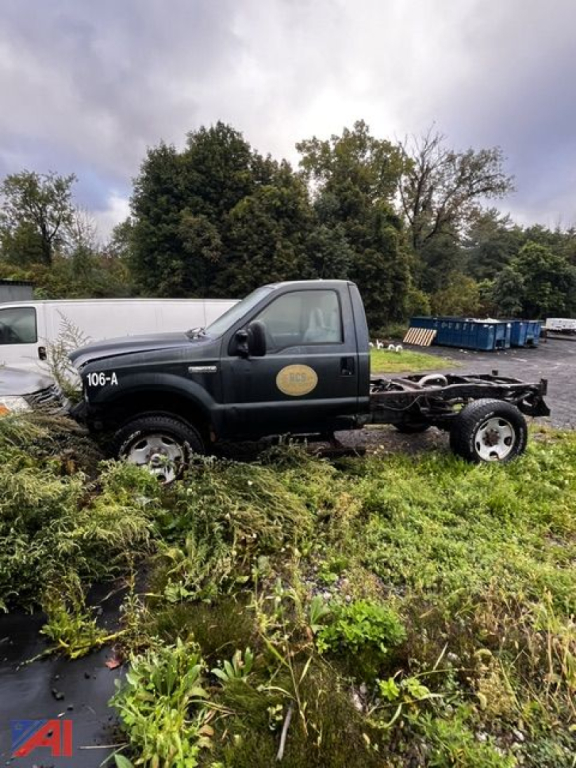 Ford F-350 cab and chassis in a field with vegetation, single rear wheel missing, cab still intact with dark green color and a yellow circle decal on the door. Visible text includes '106-A' on the cab and a circular yellow emblem. Overcast 