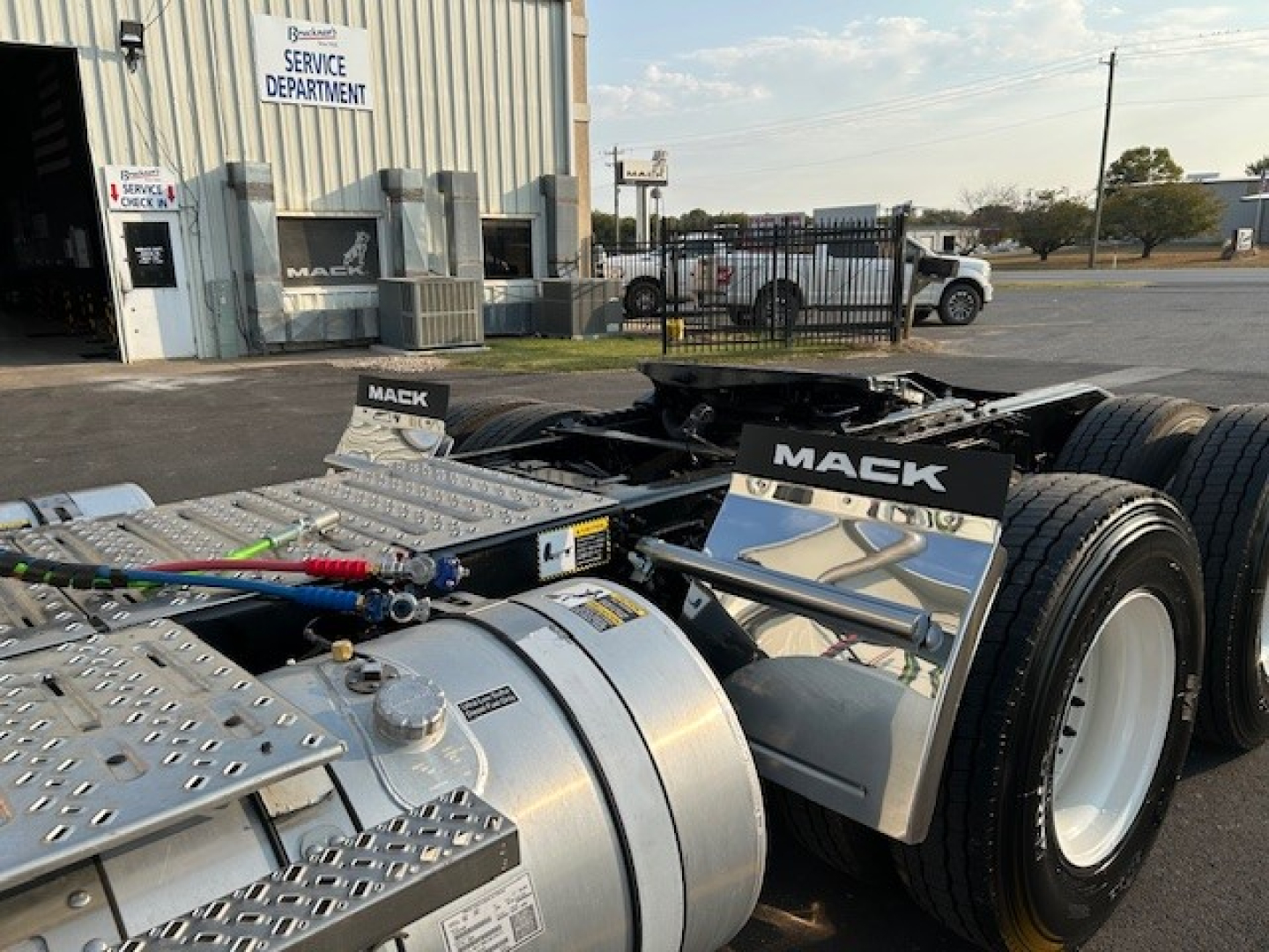 Mack AN64T truck chassis showing dual rear axles, fuel tank, and steps with stainless diamond plate; branding panels read MACK; surrounding service department building in background.